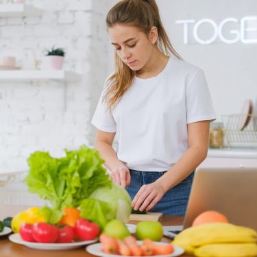 Woman in white t-shirt preparing fresh vegetables in a bright kitchen with a laptop.